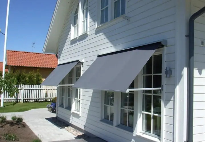Grå markiser monterade på ett vitt trähus ger skugga åt fönstren.