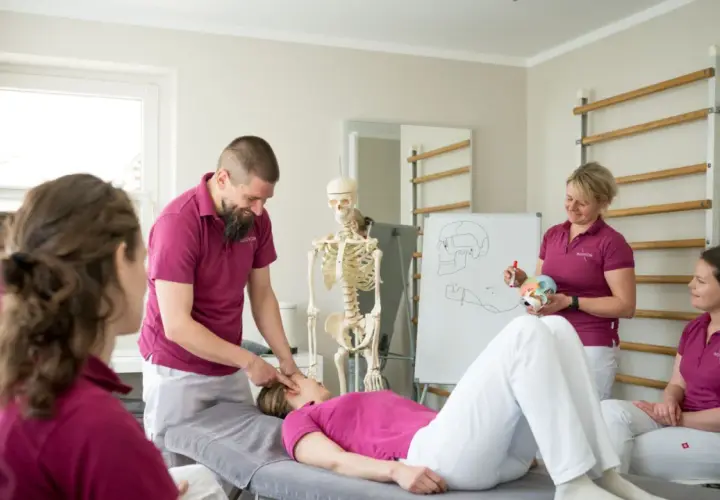 Ein Mann erklärt einer liegenden Frau eine physiotherapeutische Behandlung. Ein Skelett und eine Tafel mit anatomischen Zeichnungen verdeutlichen den Unterschied zwischen Ergo- und Physio-Therapie.