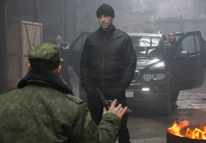 W magazynie, wśród dymu i ognia, mężczyzna w czapce i kurtce bomberce, niczym pitbull, ostatni pies, mierzy z pistoletu do żołnierza.
