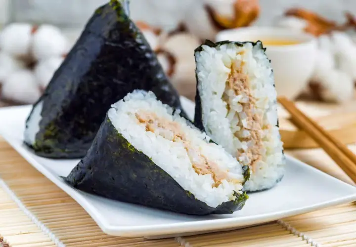 Three triangular onigiri triangle sushi with tuna filling, wrapped in nori seaweed, sit on a white plate with chopsticks and a cup of tea in the background.