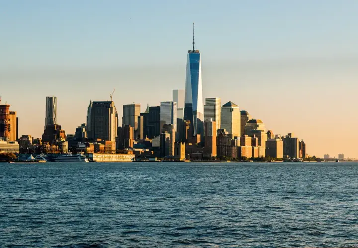 Widok na Manhattan z rzeką w pierwszym planie. W oddali widać drapacze chmur, w tym nowoczesne wieże World Trade Center, oświetlone zachodzącym słońcem.