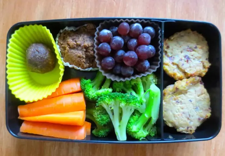 A bento box packed with bento nutrition: a date ball, muffin, grapes, carrots, broccoli, and two savory patties.