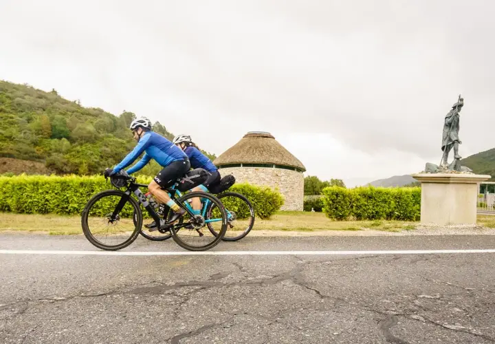 Ciclistas pedalean por la ruta EuroVelo 3, pasando junto a un edificio redondo con techo de paja y una estatua de un peregrino.