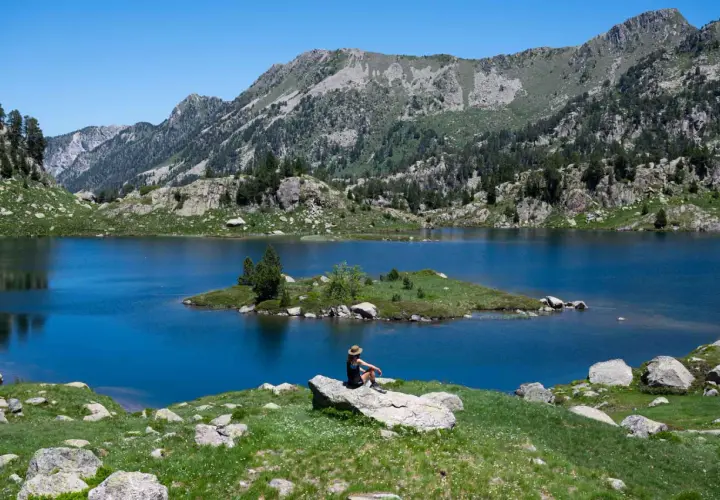 Persona sentada en roca contempla el azul intenso del circ de Colomers, rodeado de montañas verdes y un cielo despejado.