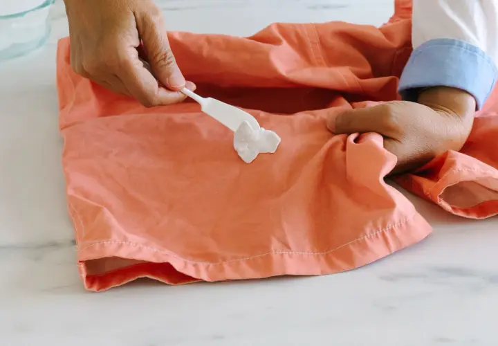 Comment enlever du chewing gum sur un tee shirt : une spatule blanche applique une pâte sur une tache de chewing gum sur un tissu corail.