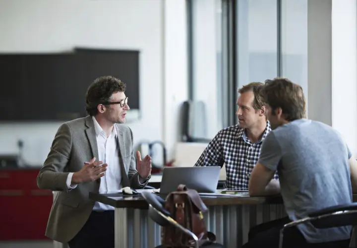 Three men discuss performance review tips for employees in a modern office setting.