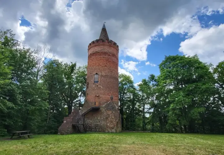Stara, ceglana wieża zamkowa z kamiennym fundamentem, otoczona zielenią drzew, pod błękitnym niebem z chmurami.