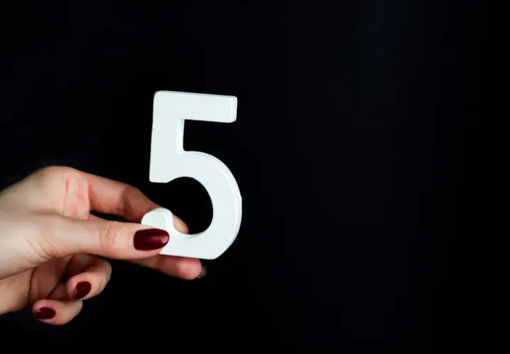 Dłoń z ciemnoczerwonym lakierem trzyma białą cyfrę 5, symbolizującą numerologię 5.
