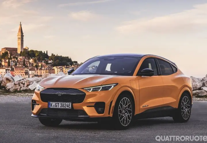 Arancione Ford Mustang Mach-E, una delle auto elettriche sportive più desiderate, parcheggiata con una pittoresca città costiera sullo sfondo.