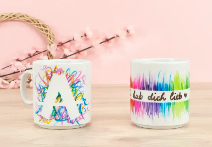 Kreative tassen bemalen ideen: eine Tasse mit dem Buchstaben A und bunte Kritzeleien, daneben eine Tasse mit Regenbogenfarben und der Aufschrift "hab dich lieb".