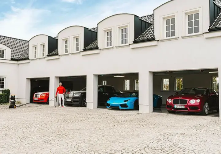 Piotr Śledź w czerwonej bluzie i białych spodniach stoi przy luksusowych samochodach: czerwonym Rolls-Royce, czarnym Rolls-Royce, niebieskim Lamborghini i czerwonym Bentleyu.