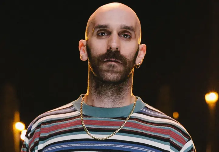 Łysy mężczyzna z zarostem, w kolorowej bluzie i złotym łańcuszku, wygląda jakby miał zaraz zacząć śpiewać "x ambassadors unsteady tekst".