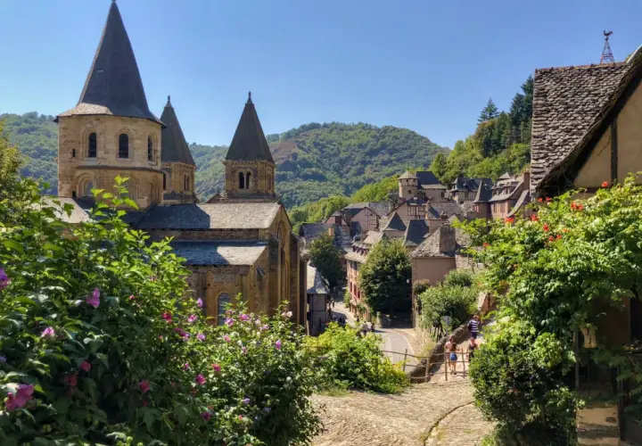 Un pueblo pintoresco con una iglesia románica y tejados de pizarra, ideal para rutas por Francia. Flores rosas y verdes exuberantes en primer plano.