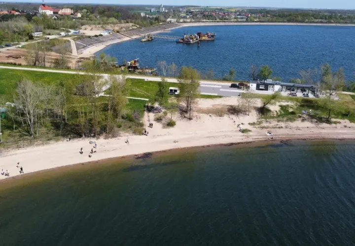 Ludzie spacerują po piaszczystej plaży nad Mietkowym zalewem. W oddali widać budynki i maszyny na wodzie.