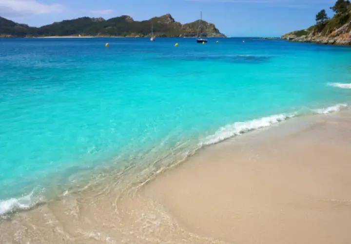 Aguas turquesas y arena dorada en unas playas cristalinas España, con veleros navegando al fondo y montañas verdes.