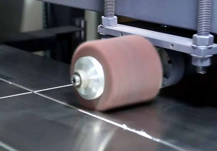 Máquina puliendo acero inoxidable. Un rodillo abrasivo gira sobre una superficie metálica, dejándola brillante.