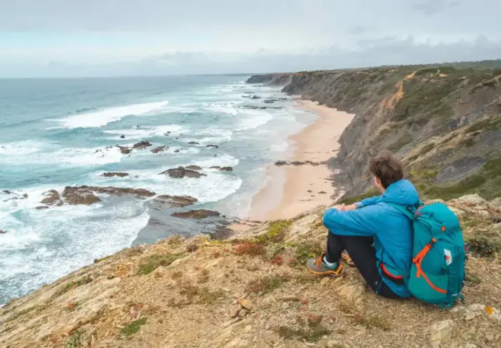 Persona con mochila azul contempla la costa salvaje durante un senderismo Portugal. Olas rompen en la playa desierta.