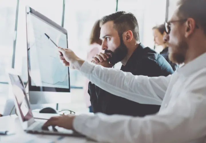Analityk biznesowy wskazuje na wykres na monitorze, omawiając dane z kolegą.