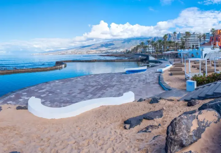 Słoneczna plaża Playa de las Américas z basenem, kolorowymi budynkami i górami w tle.