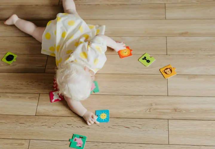 Dziecko z jasnymi włosami, w żółtej sukience w kropki, przewraca się na brzuch wśród kolorowych puzzli z zabawnymi zwierzątkami i słońcem.