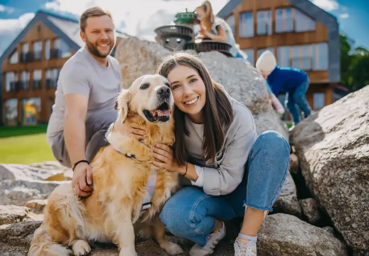 Uśmiechnięta rodzina z psem na tle drewnianych domków. Czy na Śnieżkę można z psem? Tak, jeśli jest tak szczęśliwy jak ten złoty retriever!