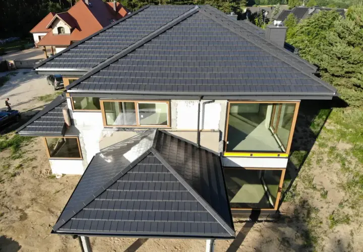 Nowy dom z ciemną dachówką, która wygląda jak blacha. Duże okna z drewnianymi ramami.