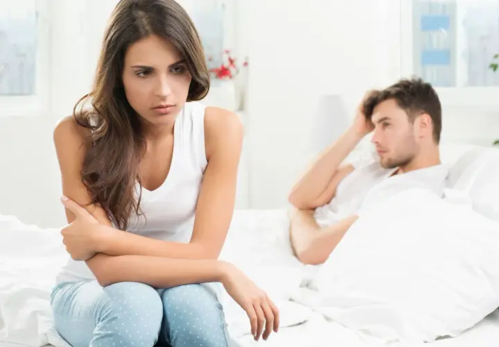 Smutna kobieta siedzi na łóżku, zmartwiona. Jej partner leży obok, z ręką na czole. Czy to uczulenie na spermę powoduje ich problemy?