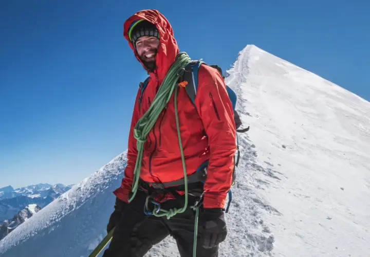 Uśmiechnięty alpinista w czerwonej kurtce na szczycie góry, z liną na ramieniu, na tle majestatycznego **Mount Blanc**.