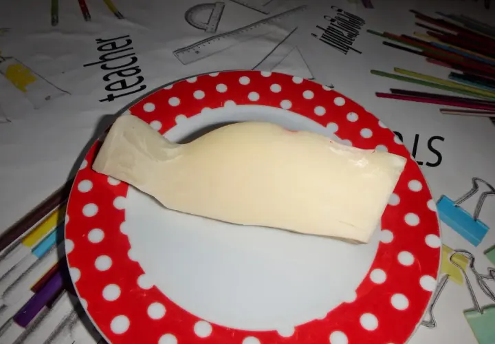 Kawałek żółtego sera provolone leży na talerzu w czerwone kropki. Tło przedstawia materiał z rysunkami przyborów szkolnych.