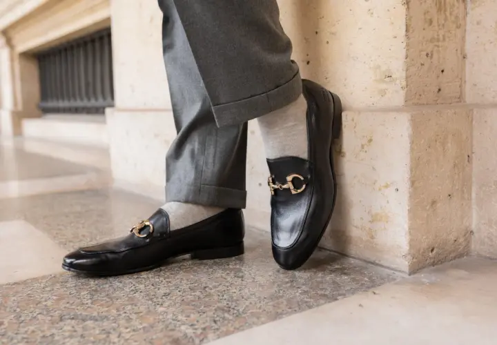 Szare, gładkie skarpetki do mokasynów z czarnymi, lakierowanymi butami i złotą klamrą. Elegancka stylizacja.