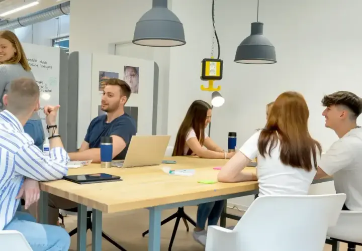 Junge Leute im **Master berufsbegleitend NRW** diskutieren an einem Tisch. Eine Frau schreibt an einer Tafel.