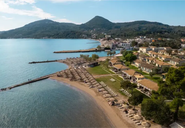 Piękna plaża z rzędami leżaków i parasoli, domki z dachówką, góry w tle. Idealne miejsce na wakacje, jak sugerują messonghi beach holiday resort opinie.