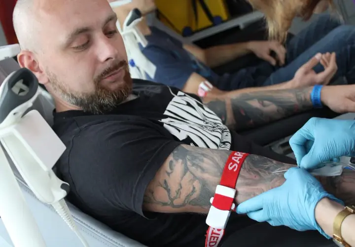 Mężczyzna z tatuażem na ramieniu oddaje krew. Czerwona opaska uciskowa i igła są widoczne.