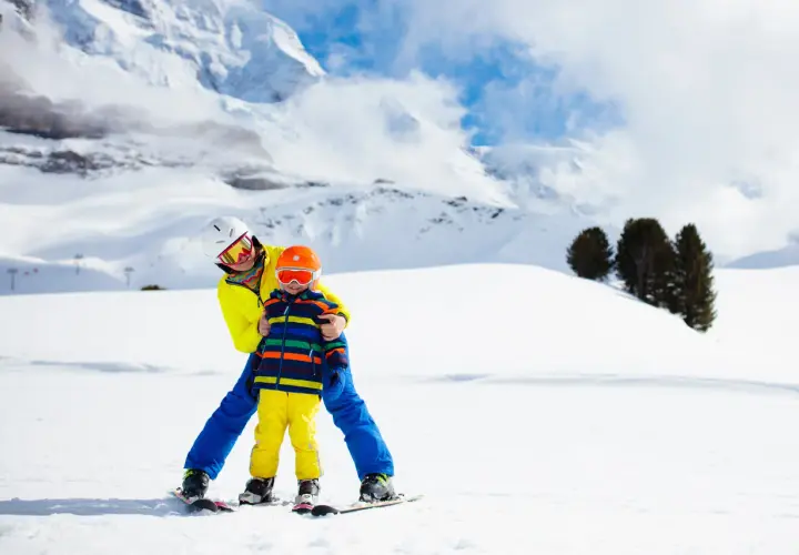 Un adulto insegna a sciare a un bambino di 2 anni su una pista innevata, con montagne sullo sfondo. Un perfetto weekend con bambini 2 anni!