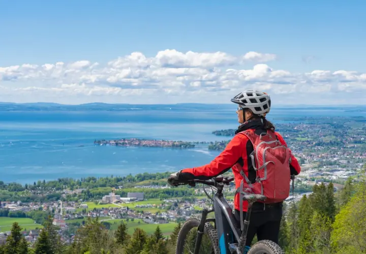 A woman in a red jacket and helmet pauses her **Lake Constance bike tour** to admire the expansive blue lake and distant towns under a cloudy sky.