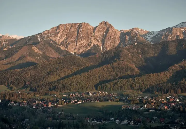 Widok z **Antałówki punkt widokowy** na Tatry. Słońce oświetla skaliste szczyty i lasy, a w dole widać zabudowania.