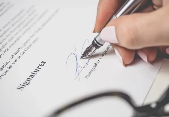 Mano con pluma estilográfica firma un documento. La firma se realiza encima o debajo del nombre "Photographer's Signature".