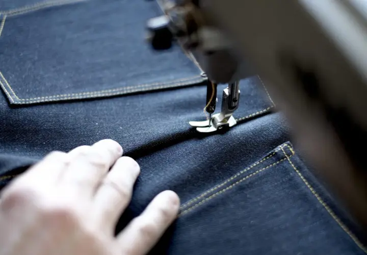A sewing machine stitches a precise jean seam on dark denim, with a hand guiding the fabric.