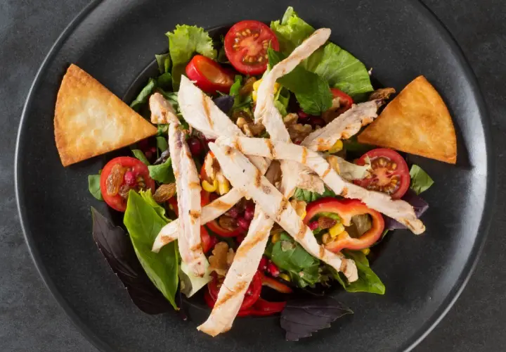 Deliziosa insalata di pollo fredda con pomodorini, mais, peperoni e crostini croccanti su un piatto scuro.