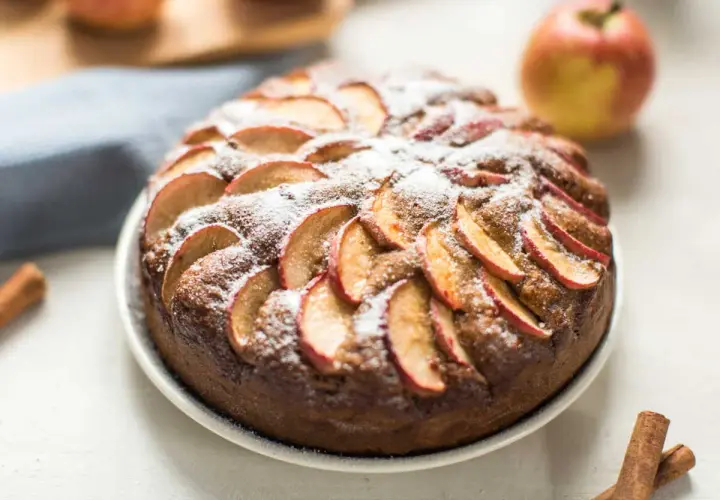 Un délicieux moelleux aux pommes, saupoudré de sucre glace, avec des pommes fraîches et des bâtons de cannelle en arrière-plan.