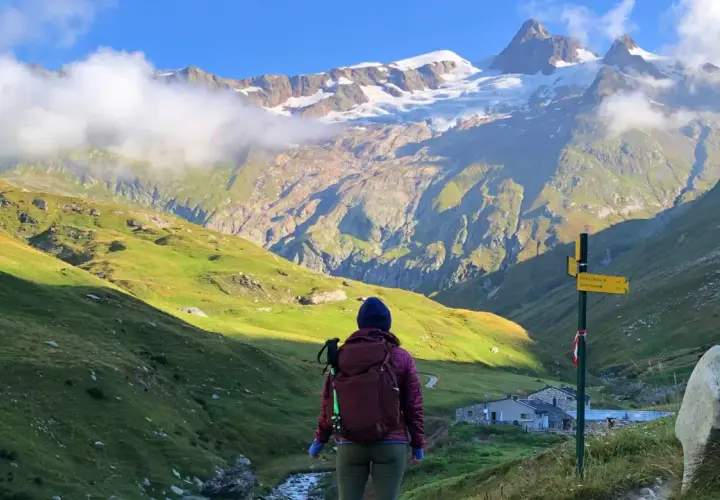 A hiker pauses on a **Tour Mont Blanc hike**, gazing at a majestic snow-capped mountain range. A signpost points the way to the next stage of this epic journey.