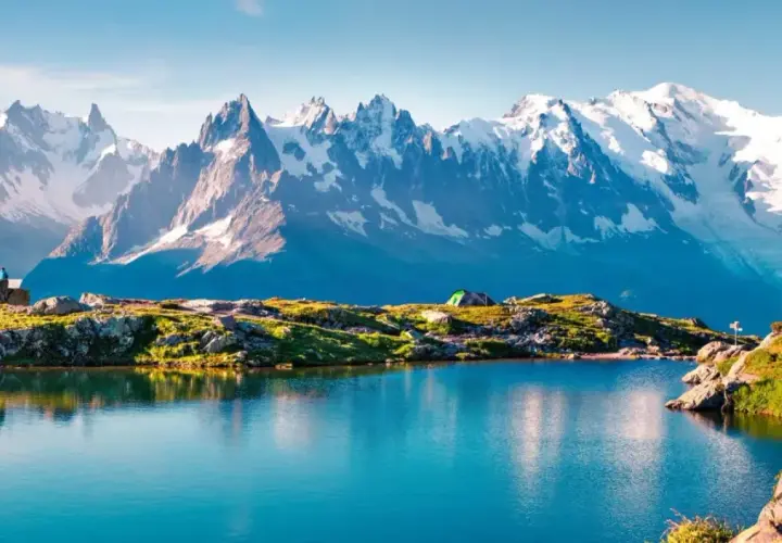 Un lago azul refleja la majestuosa **montaña Mont Blanc** y sus picos nevados. Un campista contempla el paisaje.