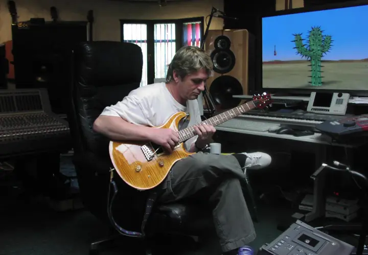 Mike Oldfield w studiu, gra na gitarze. Na ekranie komputerowym kaktus na pustyni, jak w jego utworach.