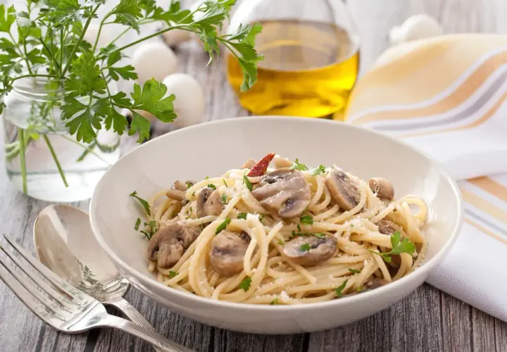 Talerz spaghetti z pieczarkami, posypany parmezanem i natką pietruszki. Obok sztućce, oliwa i zioła.