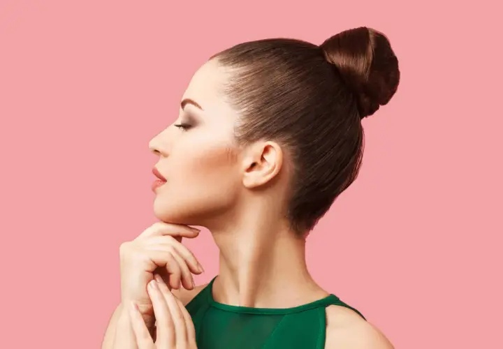 Mujer con moños altos elegantes, perfil sereno, mano en la barbilla, fondo rosa.