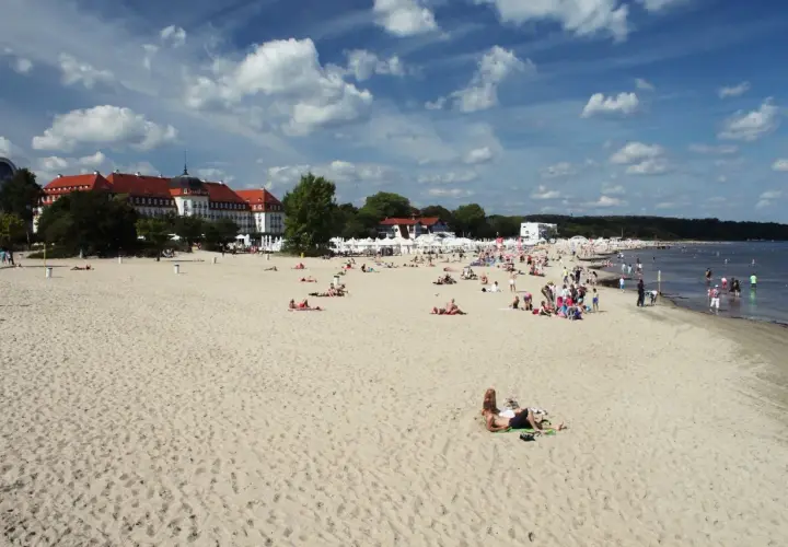 Plaża w Sopocie z ludźmi opalającymi się i spacerującymi, w tle budynki i morze.