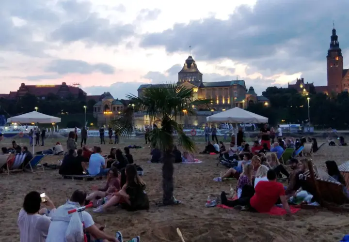 Ludzie odpoczywają na plaży, ciesząc się wieczorem. W tle widać piękne budynki, świadczące o bogactwie wydarzeń kulturalnych w Szczecinie.
