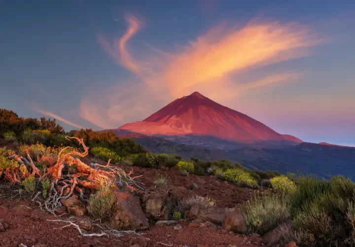 Volcán Teide bañado por el atardecer, ideal para actividades de montaña. Nubes anaranjadas y vegetación árida en primer plano.