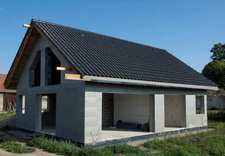Dom w stanie surowym otwartym, z dachem pokrytym czarną dachówką. Widoczne otwory okienne i drzwiowe, betonowe ściany.