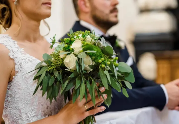Panna młoda trzyma bukiet ślubny z białymi różami i zielenią, obok pan młody w garniturze.